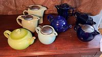 Seven assorted teapots displayed on a wooden surface including yellow-green Le Creuset and dark blue ceramic styles