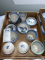 Box containing a set of M.A. Hadley pottery including various cups, small plates, and salt and pepper shakers with blue hand-painted folk art style decoration.