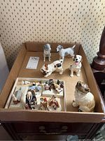 Overview of the lot including four dog figurines, cat figurine, and miniature indoor scene figurines arranged in cardboard box on wooden table.