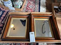 Two rectangular wooden framed mirrors placed side by side on a table with a numbered tag between them, showing their wood grain and gold accent trim details.