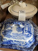 Two ceramic serving dishes: a cream-colored Spode Italian lidded server with white rabbit lid handle showing crazing and chip, alongside a blue and white ceramic lidded serving dish with pastoral scene and decorative handles.
