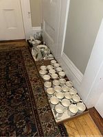 Long view of the lot showing multiple trays of dinnerware and serveware arranged in a hallway, includes stacked plates, cups and various serving dishes.