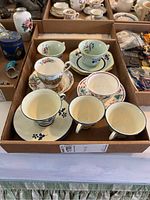 Seven assorted teacups and saucers in a cardboard tray showing variety of floral and pastel designs.