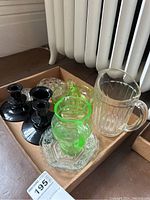 Glass pitcher, black candlestick holders, green glass citrus juicer, glass flower frogs, and clear pressed glass plate arranged in a box.