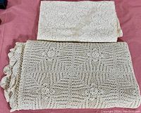 Two folded antique linens, one crocheted with a floral motif and scalloped edges, the other lace with floral pattern, displayed on a red surface.