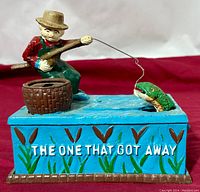 Front view showing the fisherman with his fishing rod, the basket, the blue base with water details, and the inscription "THE ONE THAT GOT AWAY."