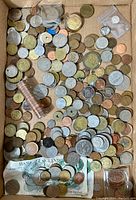 Wide overhead image showing a large assortment of old coins loose in a cardboard box with some visible paper currency at the bottom.