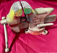 Heavy duty used steel bench vise on red surface showing the front and side profiles including the handle and locking lever.