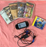 Sony PSP console powered on with screen visible, six UMD game and video cases including Dragonball Z, Avatar, Sid Meier's Pirates, Need for Speed, The Exorcism of Emily Rose, and Pokémon movie, with AC adapter connected.