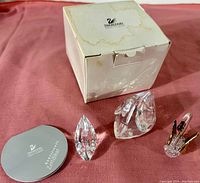 Photo showing three Swarovski crystal swan figurines alongside original box and certificate of authenticity on a pink cloth.