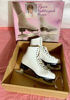 Pair of white figure skates inside original box with a visible image of the packaging showing figure skates and a woman.