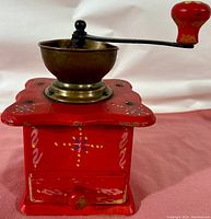 Side view of red wooden coffee grinder with hand-painted folk art designs, steel handle with red knob, and brass grinding hopper embossed with DE logo.