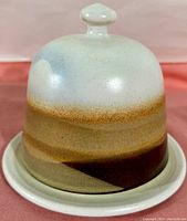 Front view of tall rounded dome lid with knob handle, showing horizontal bands of warm browns and cool gray-blue glaze.