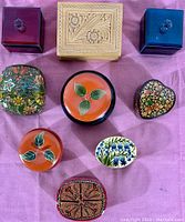 Overhead view of nine vintage trinket boxes on pink fabric, showing shapes, materials, and painted floral designs.
