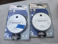 Two Kidde brand carbon monoxide alarms in original packaging, each with attached battery, shown on a flat gray surface.