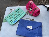 Photo of three purses on a white table: pink quilted chain shoulder bag, turquoise studded clutch with tag, and blue envelope clutch with gold-tone twist lock.