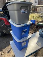 Photo showing three stacked plastic storage totes: one gray and two blue with white lids, situated outdoors near a car and table.
