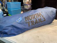 Blue fabric bag with black North Trail logo and model name Outpost 1000 III containing the tent, placed on a surface indoors.