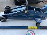 Side view of blue hydraulic trolley jack with detached handle and pressure gauge hose on white table surface