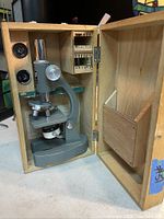 Open wooden case displaying the gray metal microscope inside, showcasing the nosepiece, eyepiece, and stage.