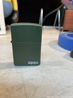 Green Zippo lighter placed on a light-colored surface, showing front view with white Zippo logo on bottom right corner.