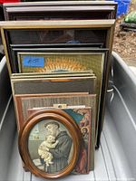 Photo showing a collection of several picture frames stacked in a plastic bin, including an oval wooden frame with needlepoint art of a saint holding a child