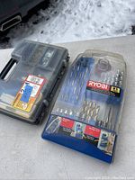 View of two plastic cases containing Ryobi drill bits and accessories including drill bits, hole saws, screwdriver bits, and other hardware-related bits laid out on a gray surface outdoors.