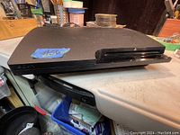 Top and angled view of black Sony PS3 console showing surface wear and a blue sticker with lot number.