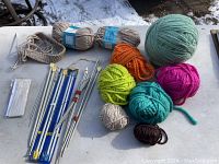 Yarn balls and skeins laid out with knitting needles and accessories visible on outdoor surface with snow.