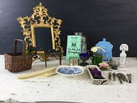 Wide shot of collection featuring ornate gold/brass cast iron frame, blue ceramic house, glass jar, silver salt cellar, wooden basket, decorative plate, and small figurines and collectibles
