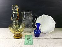 Full view of all five glass art pieces together on white surface against dark background showing owl figurine, amber bowl, clear etched pitcher, cobalt mini pitcher, and white rose bowl.