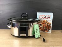Front view of stainless steel Crock-Pot slow cooker with glass lid and cookbook standing behind it.