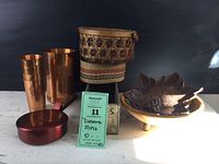 Photo showing the copper pitcher, four copper glasses, two small woven baskets, footed wooden bowl, wooden cheese box, and grape leaf metal dish all arranged on a light surface against a black backdrop.