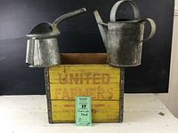 Photo showing front view of the crate with the two galvanized containers sitting inside. Crate is yellow with faded red text 'UNITED FARMERS'. The watering can and oil can are visibly aged and galvanized.