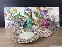 Three MacKenzie Childs cork backed placemats with colorful floral and bird patterns, one placemat shows a scratch, displayed with two floral plates, a hurricane vase, and a clear glass candelabra with dangling crystals.