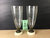 Pair of hurricane candleholders shown from front. Both have clear glass globes with etched floral patterns, white marble bases, and metal holders.