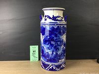 Full front view of the tall blue and white ceramic vase with floral design and decorative handles, placed on a wood surface against a black background.