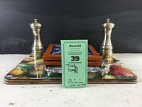 Two Old Thompson silver tone salt and pepper grinders in front of four fruit placemats, two Greek tile trivets, and Arthur Court cheese board stacked behind.