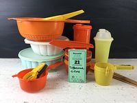 Collection of assorted Tupperware and kitchen items including a light blue covered jello mold, an orange strainer, small measuring cups, silicone scrapers, and bitters cups with handles.