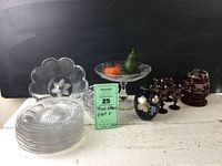 Various vintage clear and red glassware displayed on a table showing plates, bowls, cordials, decorative fruits, and a black vase.