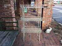 Front view of the three-tier wire shelves placed outdoors near brick pillars. Shows full structure and leg design.