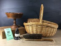 Photo showing collection of wood and wicker items: large oval wicker basket with handle, smaller basket, wood compote on pedestal, shabby wood box, black cast iron twine holder, and dark bristle dust brush with wooden handle.