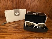 Photo showing off white beige wallet with textured flap and metallic twist lock next to white framed Callaway sunglasses in black branded case