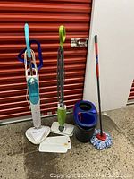 Photo showing two steam mops, a mop pail with wringer, and a mop with handle and mop head, all on concrete ground with red door background.