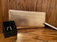 Rose gold Ted Baker wristlet clutch alongside the earrings in a black box, showing the exterior texture and brand name.