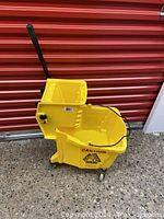 Side view of yellow commercial mop bucket with wringer and handle, showing caution label and wheels.