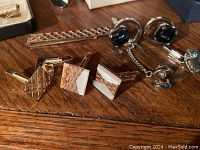 Cufflinks and tie clips arranged on wooden surface, showing variety of designs and materials including metallic finishes and stones.