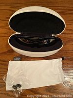 Black Oakley sunglasses inside a white hard shell case with a white cleaning pouch and a small plastic bag with replacement accessories on a wooden surface.