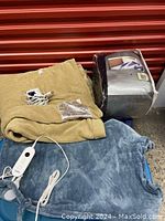 Tan and blue electric heating pads laid out with cords and controls visible, along with a navy blue blanket in a plastic bag nearby