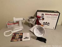 KitchenAid Fresh Prep Slicer/Shredder parts and accessories arranged on table including attachment base, food pusher, knives, booklet, and product box